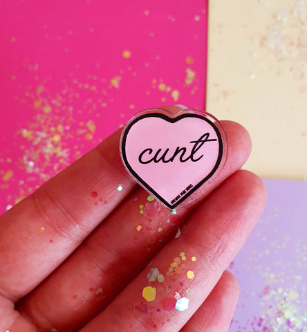 A pink heart shaped pin with the word cunt in black cursive font is being help in a hand covered in glitter. The background is colourful and glittery.